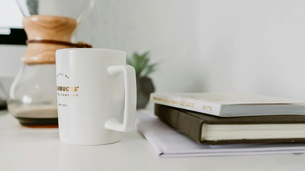 A minimalist desk with a white coffee mug, a stack of books, and a notebook, ready for a focused reading session.