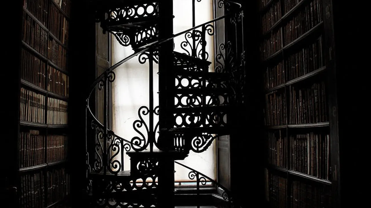 Ornate wrought-iron spiral staircase between tall shelves of old books in a dim library.