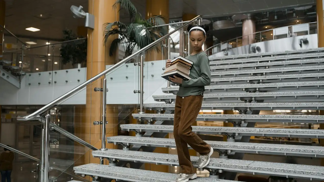 A child wearing a green sweater and brown pants with a white headband walks down metal stairs in a modern library, carrying a stack of books.