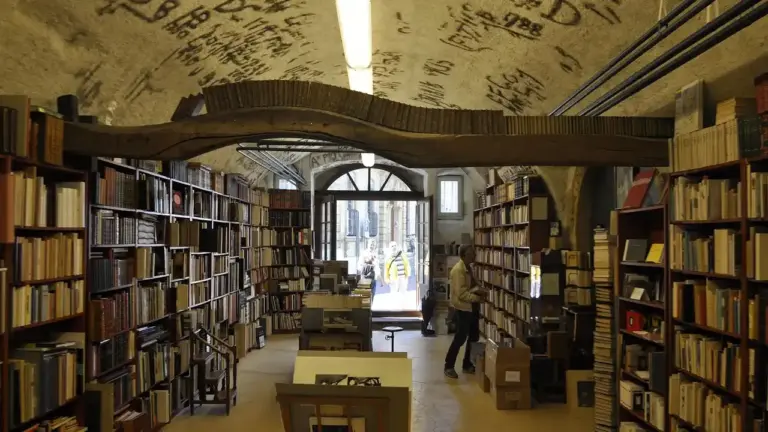 Interior of a library or bookstore with tall shelves filled with books and a few people browsing.