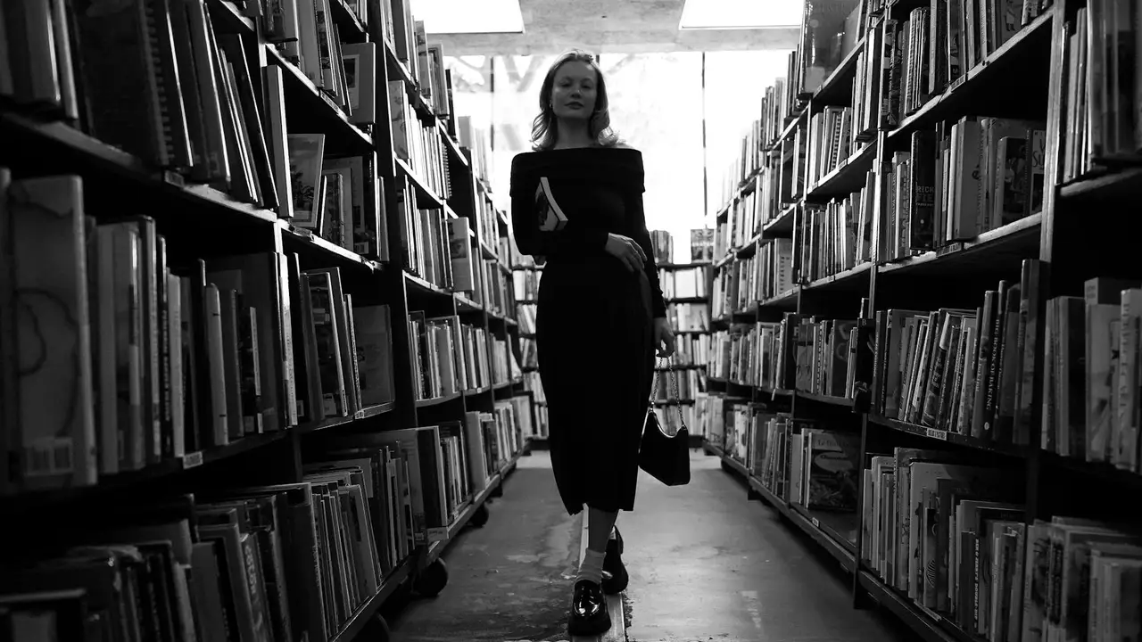 In black and white, a woman in a dark dress walks down a library aisle between tall shelves, holding a book as light streams overhead.