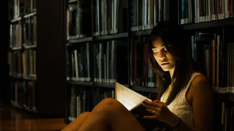 Young woman sitting among tall library shelves, absorbed in a book under warm, ambient lighting