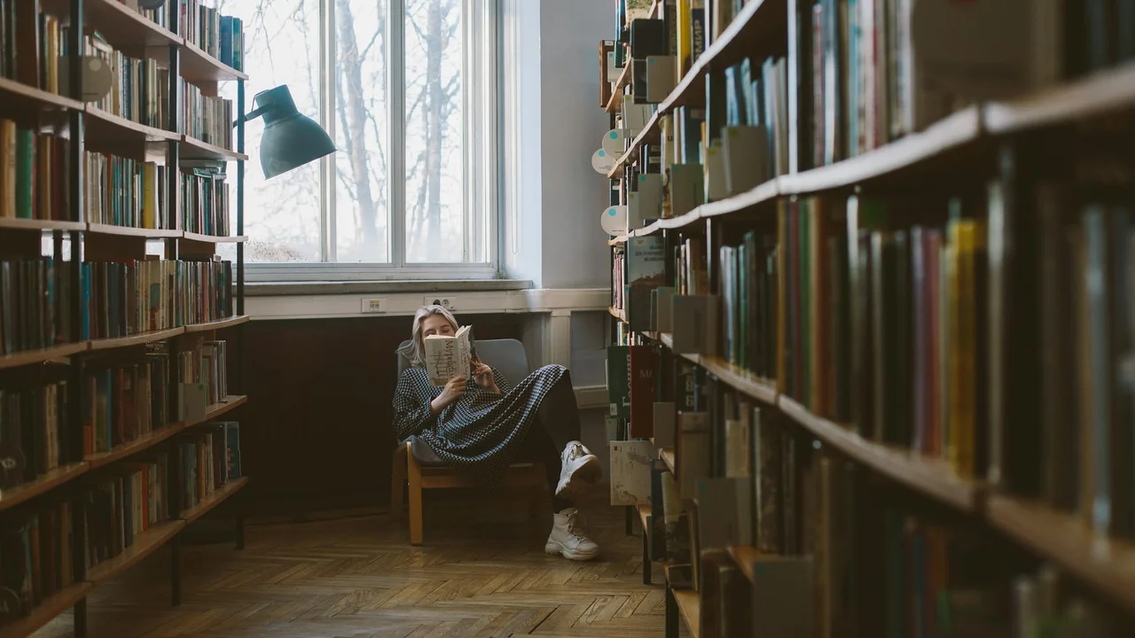 A person sits in a quiet library reading a book, flanked by tall shelves of books with a window behind.