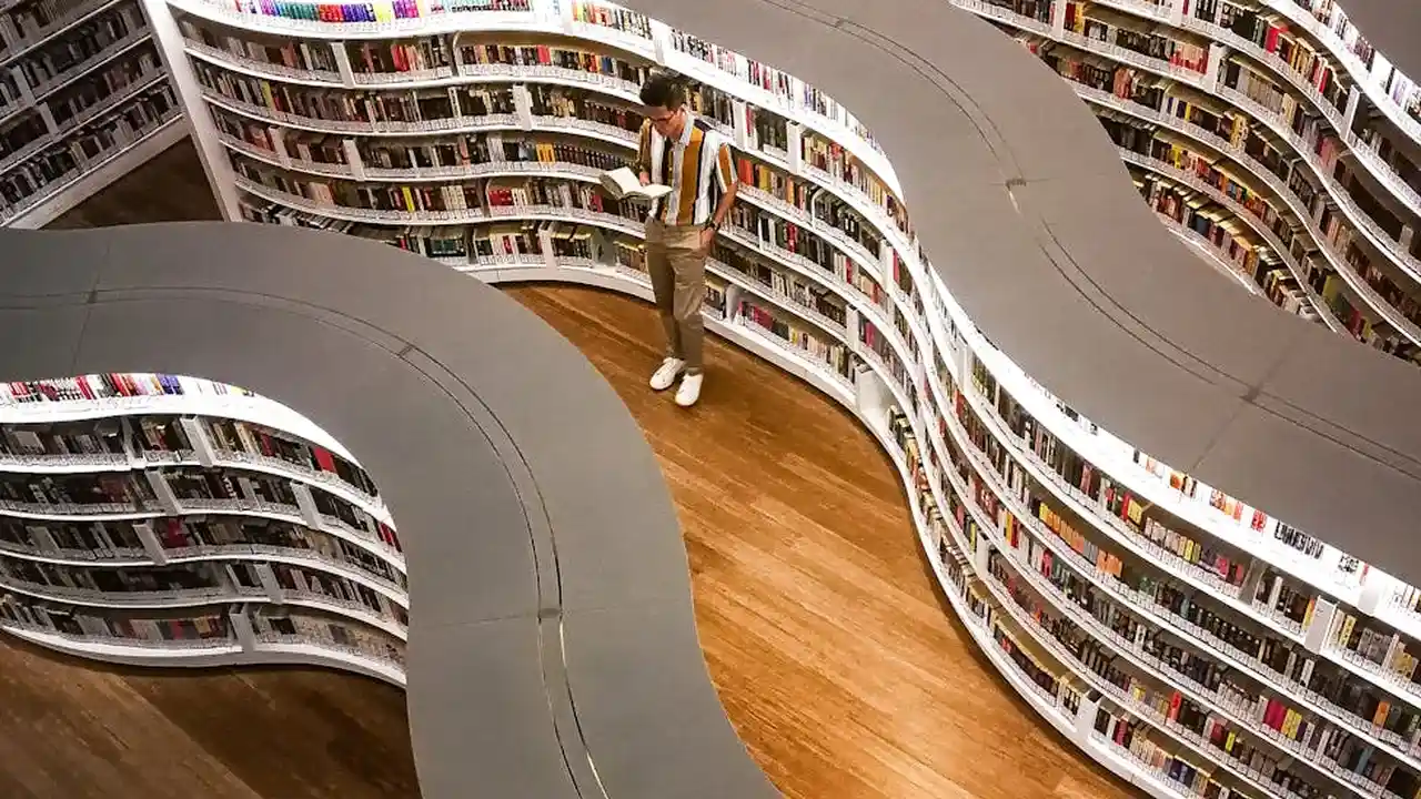 A person stands on a wooden floor in a modern library with white, curved, spiral shelves filled with books.