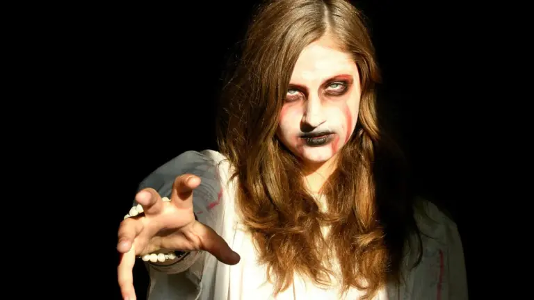 A person with pale makeup resembling a ghost or zombie, reaching toward the camera in a dark background.
