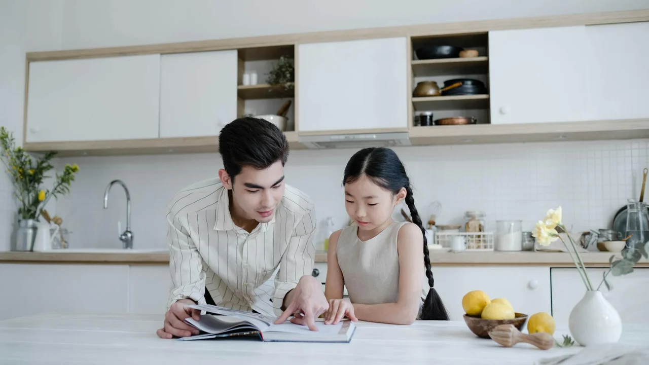 A man and a young girl sit at a bright, modern kitchen counter looking over an open cookbook together.