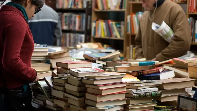 Inside a busy bookstore with piles of books on tables and shelves in the background, customers browse among the stacks.