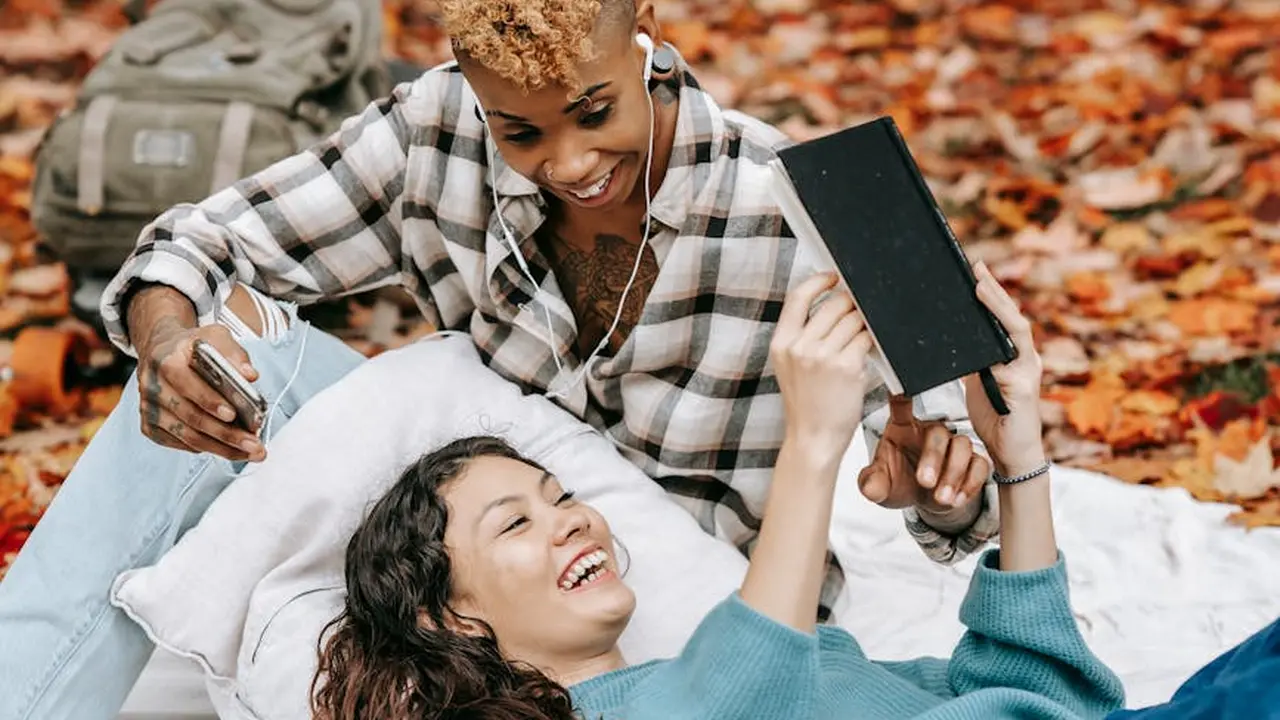 Two people lying on a blanket in autumn leaves, smiling and sharing a book and a tablet.