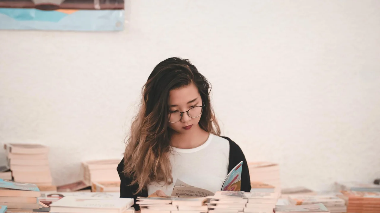 A young woman with long hair and glasses sits at a table surrounded by stacks of books, reading a paperback.