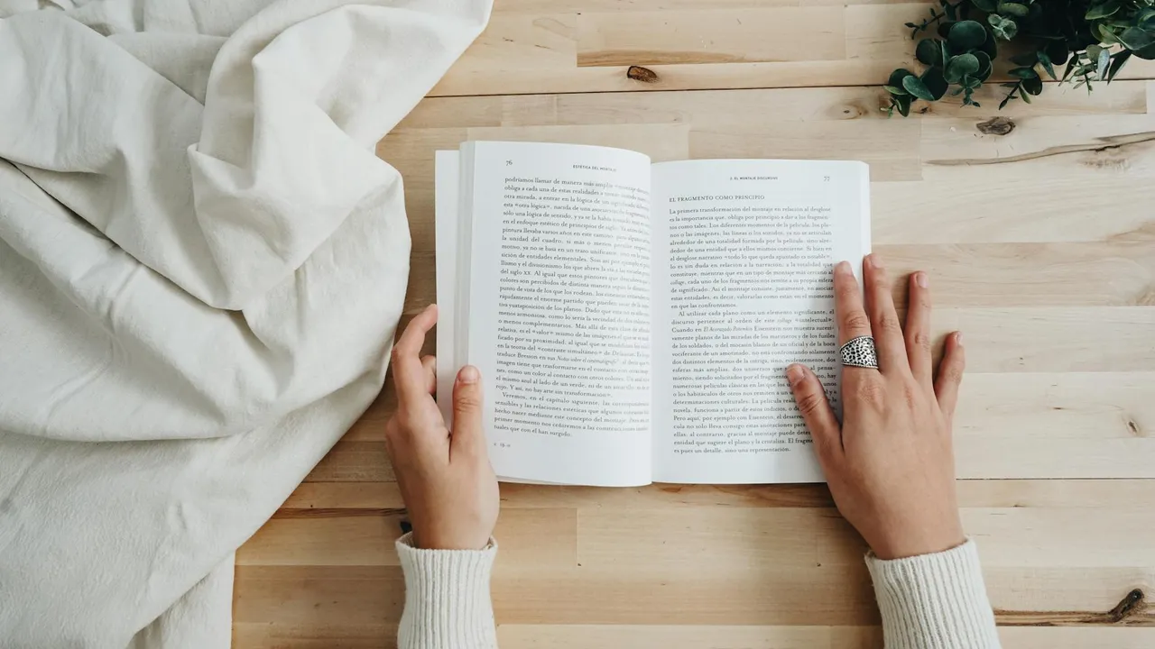 Close-up of a person reading an open book on a light wooden table; hands in a white sweater, with a white blanket on the left and a small plant in the top right.