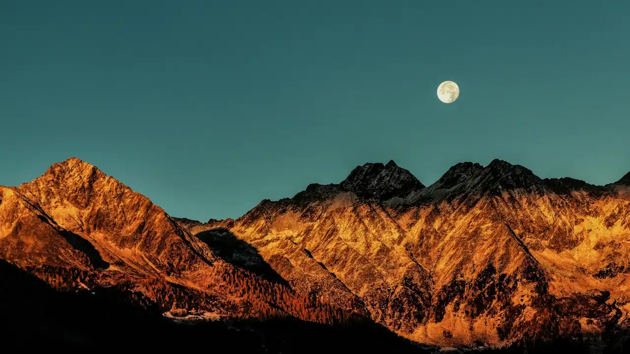 Moon rising above a rugged mountain range bathed in warm sunset light