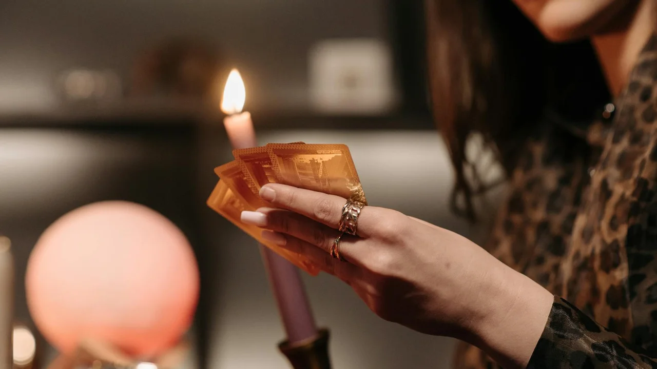 Person holding a fan of cards near a lit candle in a softly lit room, creating a dreamy, mysterious nighttime mood.