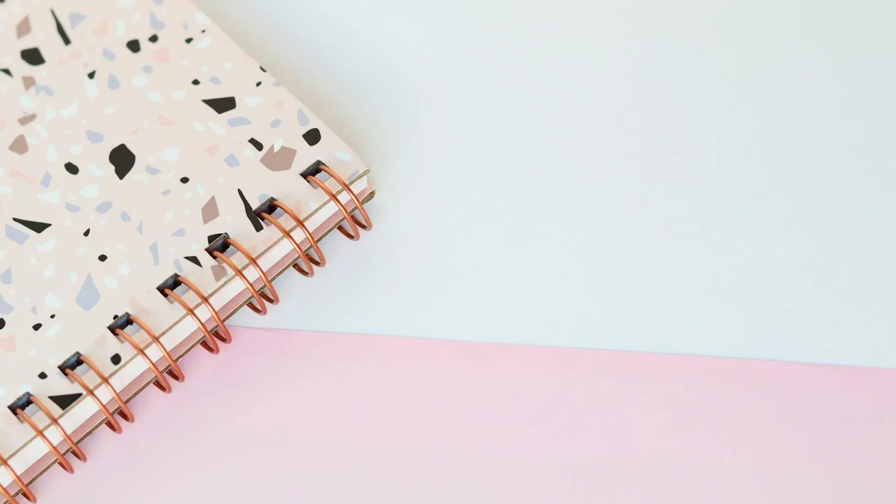 Close-up of a terrazzo-patterned notebook with copper spiral binding beside a pastel pink notebook on a light blue surface.