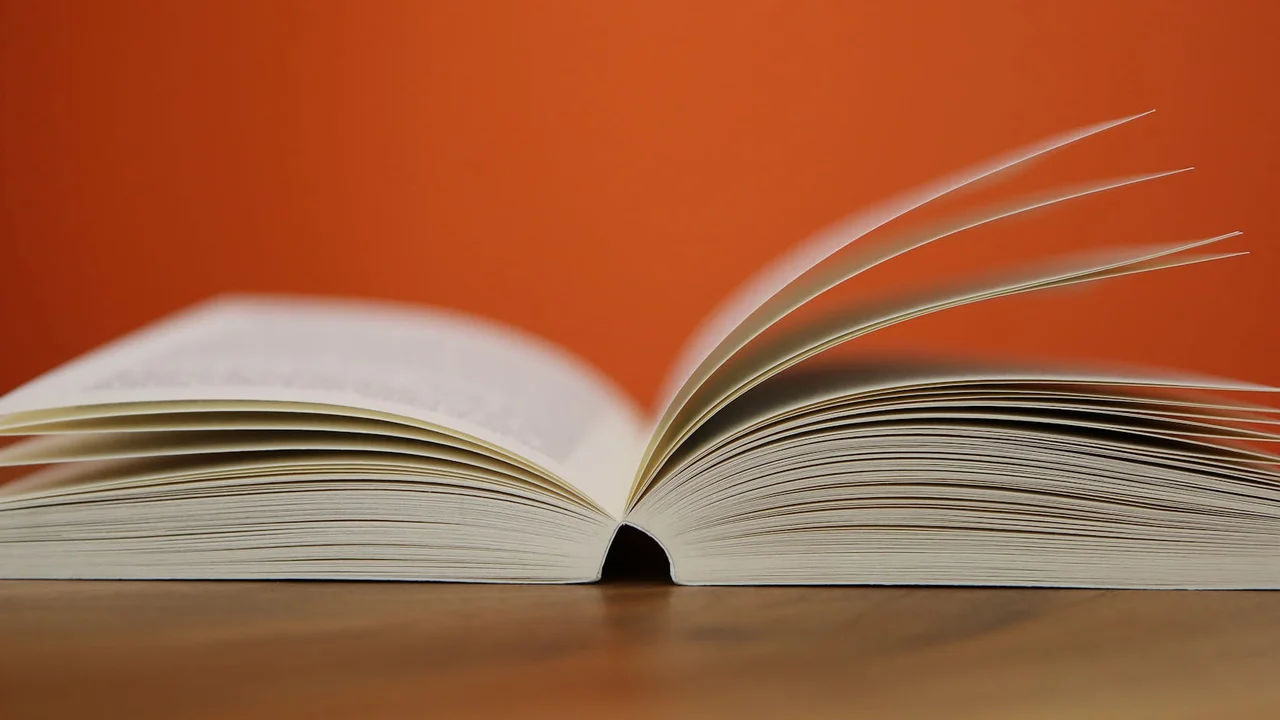 Close-up of an open book laid flat on a wooden surface with pages gently fanned, against a warm orange background