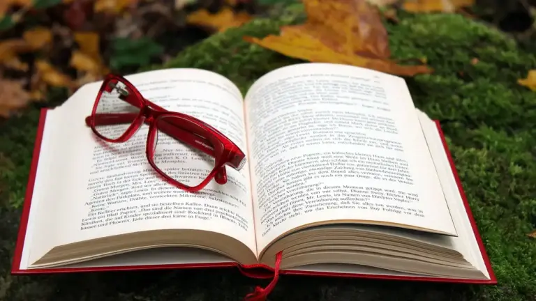 Open book with red-framed glasses resting on its pages, set on green moss with autumn leaves in the background.