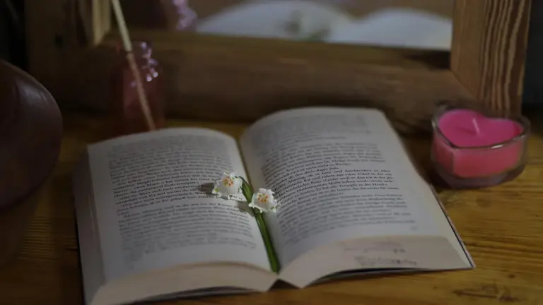An open book on a wooden table with a pink candle and a small glass nearby, evoking a cozy reading setup.