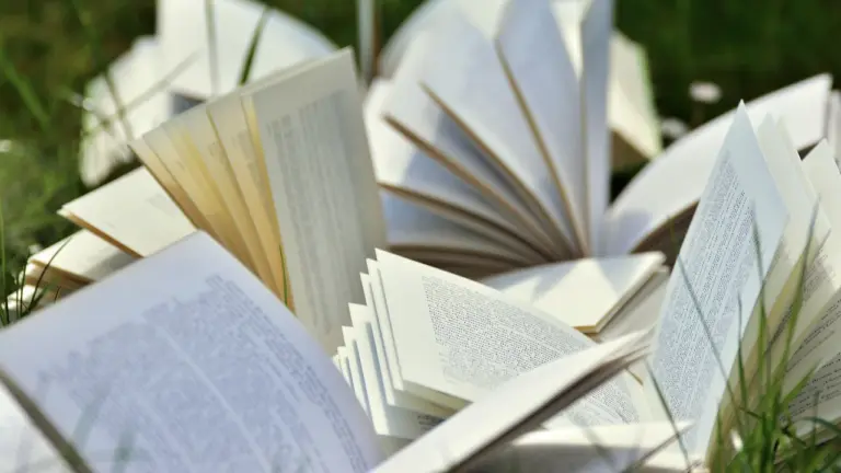 A collection of open books spread across green grass, with pages fanned and fluttering in the breeze.
