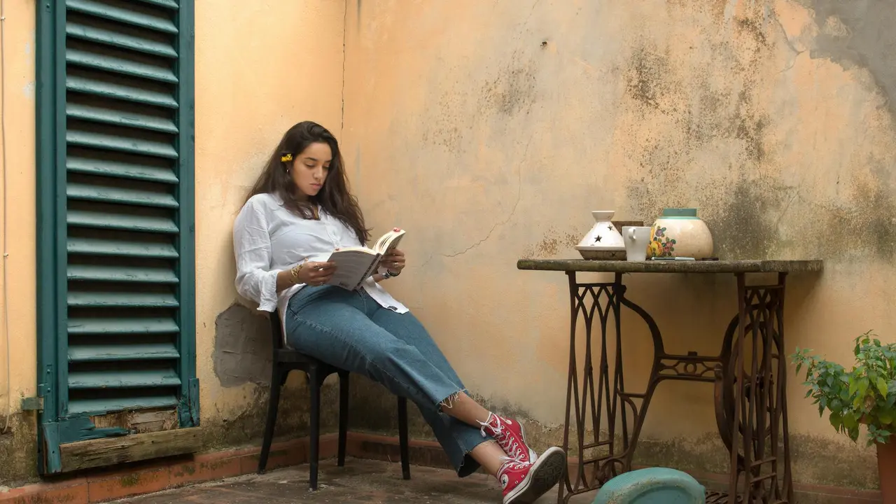A person sits on a chair in a sunlit outdoor courtyard, reading a book, with a small table, potted plants, and a weathered wall in the background.