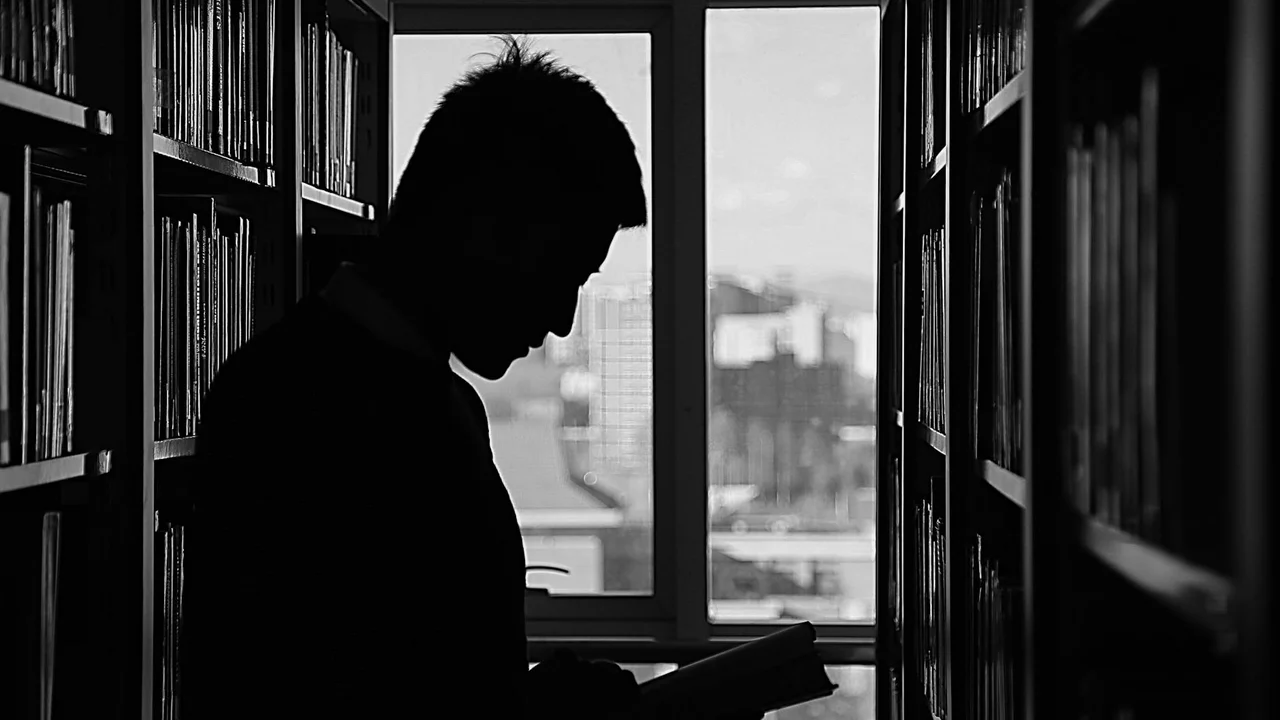 A silhouette of a person standing in a narrow library aisle, surrounded by tall shelves and looking down at a notebook.