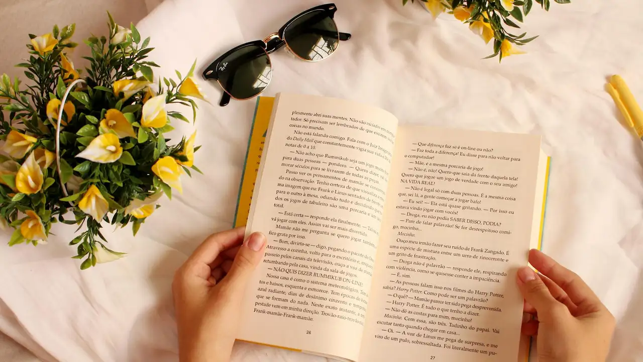 Hands holding an open paperback on a white surface, with a bouquet of yellow flowers and a pair of sunglasses nearby.