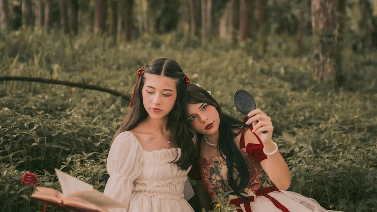 Two young women in vintage-style dresses sit in a forest; one reads a book while the other looks into a handheld mirror.