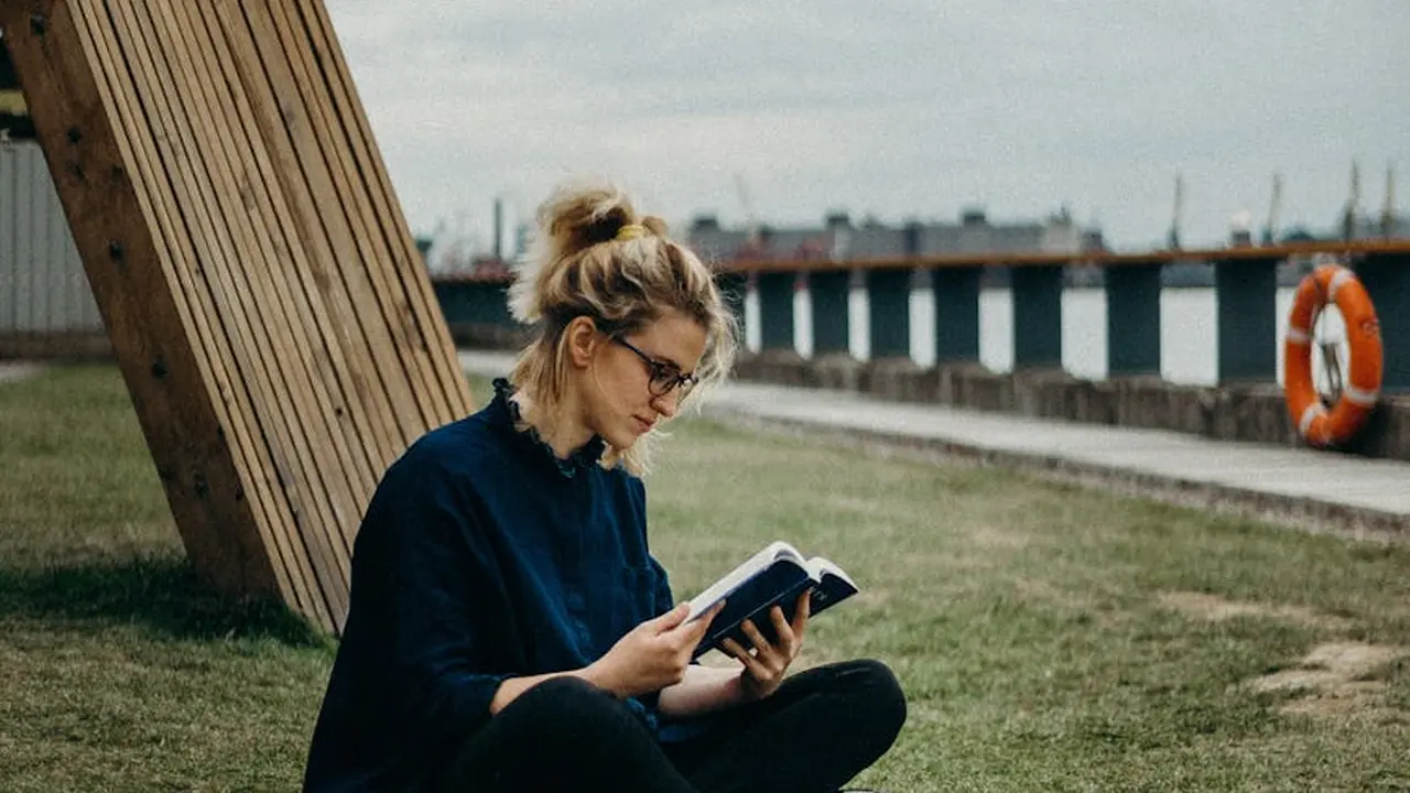 A woman with glasses sits on the grass by a waterfront, reading a book, with a pier and lifebuoy visible in the background.