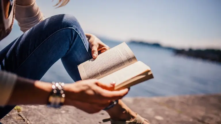 Person sitting by a lake, wearing jeans and bracelets, reading an open book with water in the background.