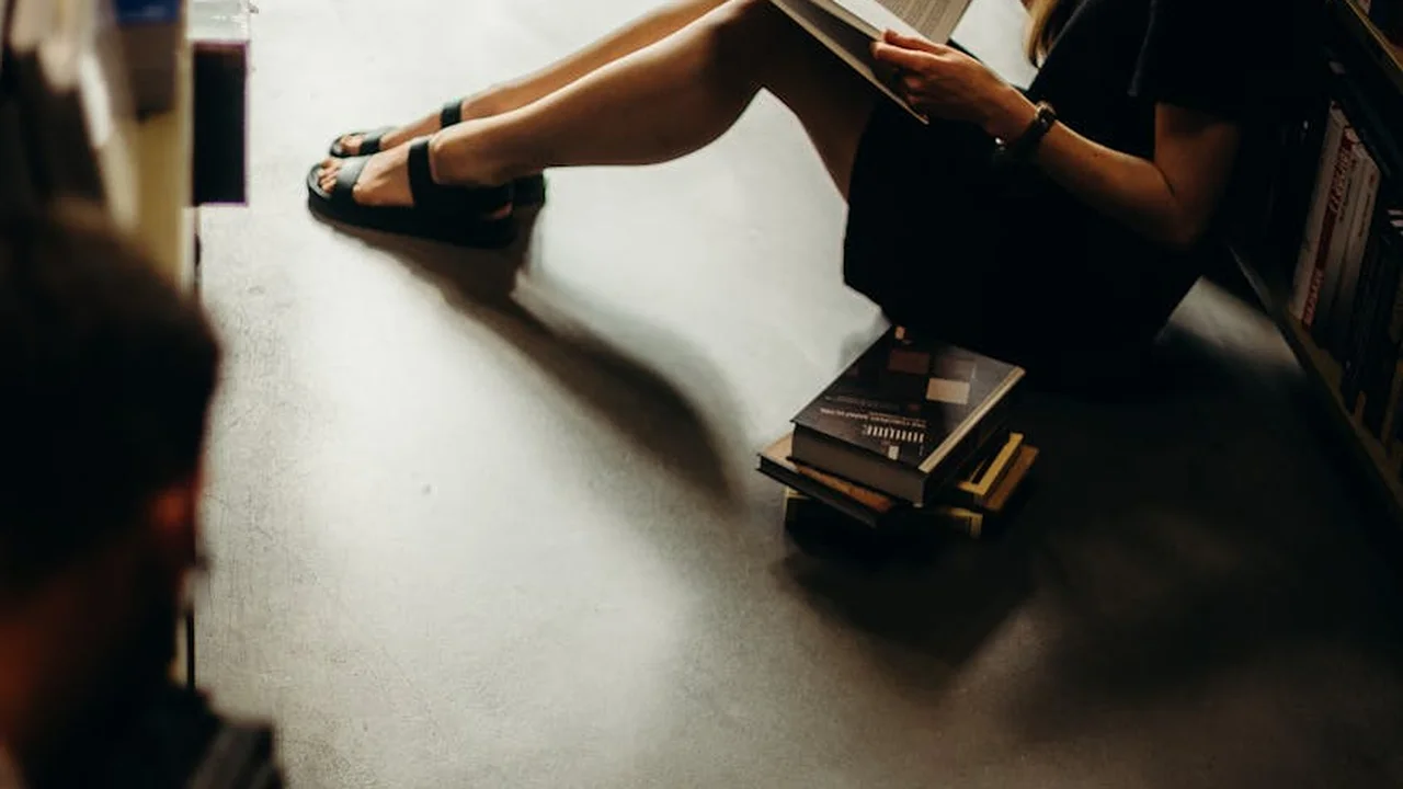 Person sitting on the floor wearing sandals, reading a book, with a small stack of books nearby.