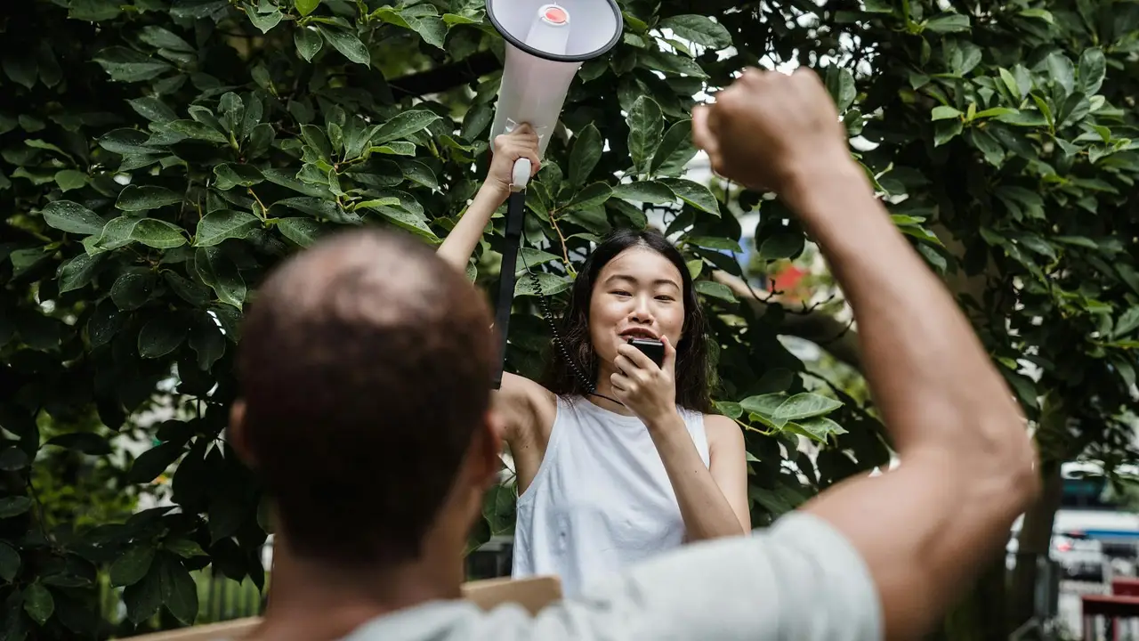 A person speaks into a megaphone amid a leafy backdrop while another participant raises a clenched fist in the foreground, evoking a protest scene.