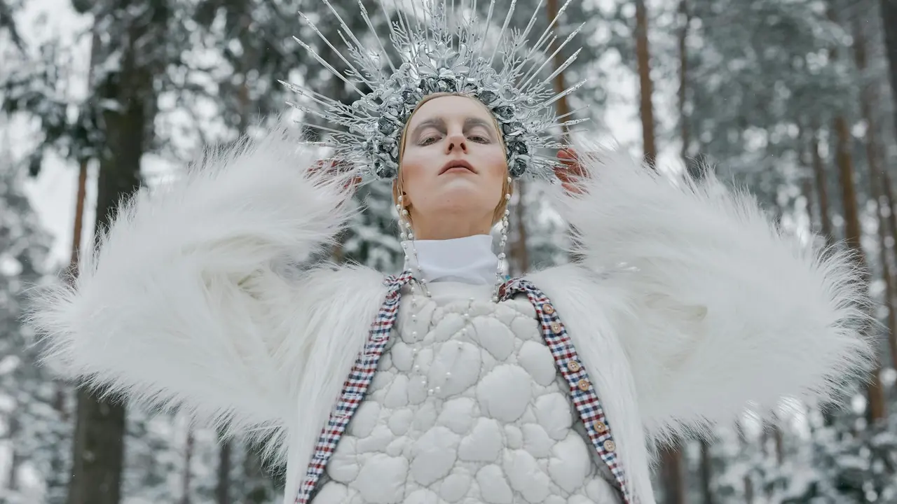 A woman wearing a white fur cloak and a spiked silver crown stands with arms raised in a snowy forest, evoking a modern fairy-tale queen.