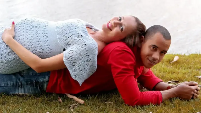 Couple lying on the grass by a lake, smiling at the camera; the woman wears a light blue knit top and the man a red shirt.