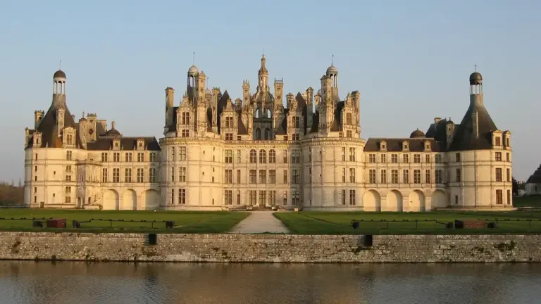 A grand French château with multiple towers sits along a river, bathed in warm light, suggesting opulent aristocratic life and courtly intrigue.