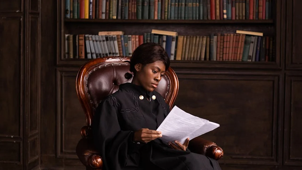 A person in a black academic gown sits in a leather armchair in a study library, poring over a sheet of paper.