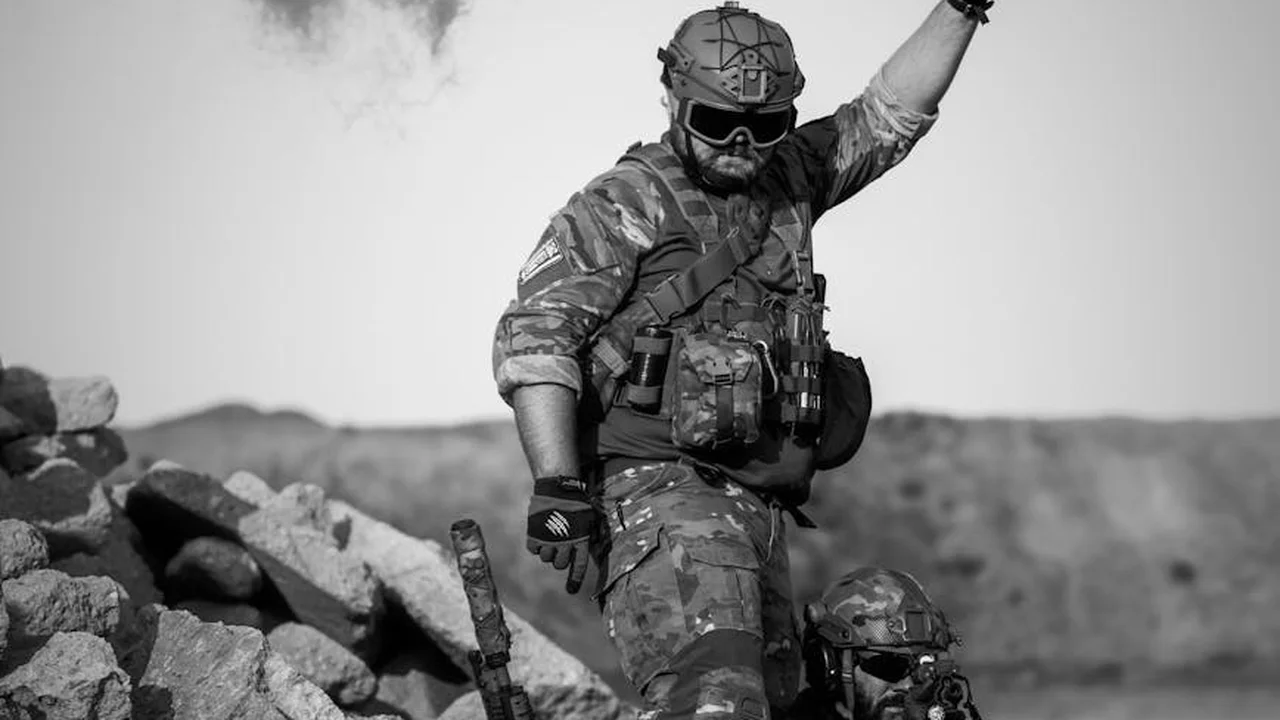 Black-and-white photo of a soldier in full combat gear standing on a rocky battlefield, helmet and goggles, with one arm raised.