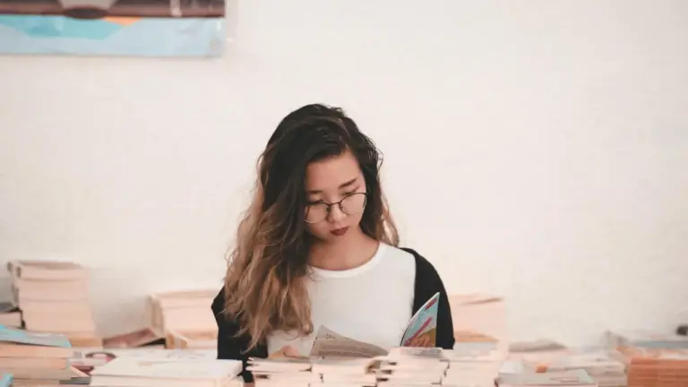 Young woman with glasses reads a book at a desk cluttered with stacked books.