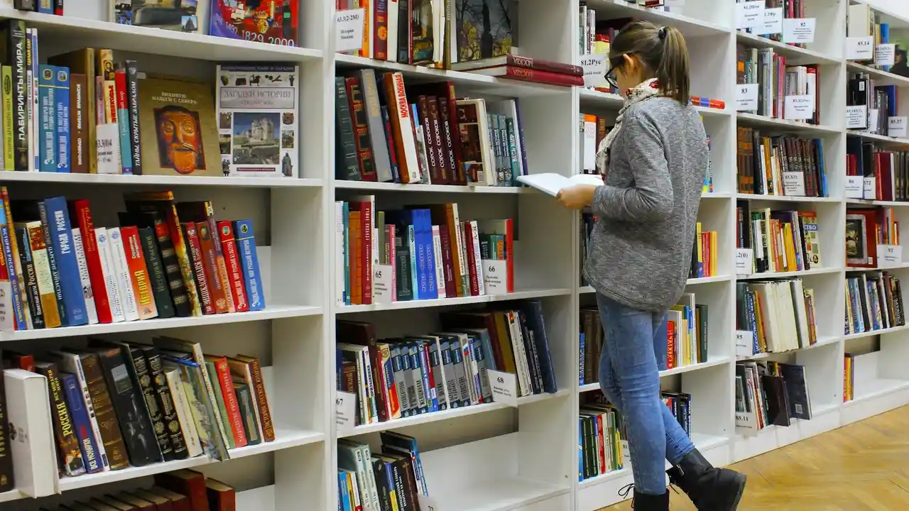 A person in a gray sweater and jeans browses a bookshelf in a bookstore or library, reading a book.