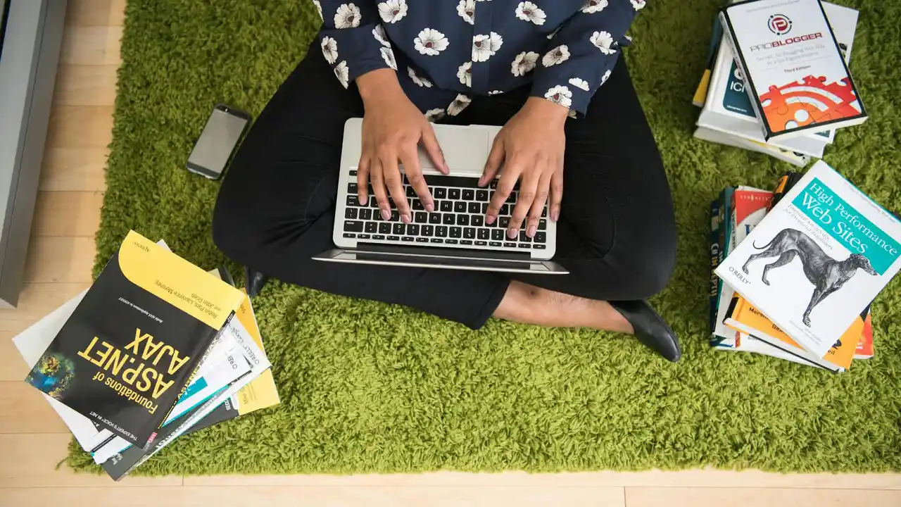 Person sits cross-legged on a bright green rug, typing on a laptop with a smartphone nearby, surrounded by piles of books.