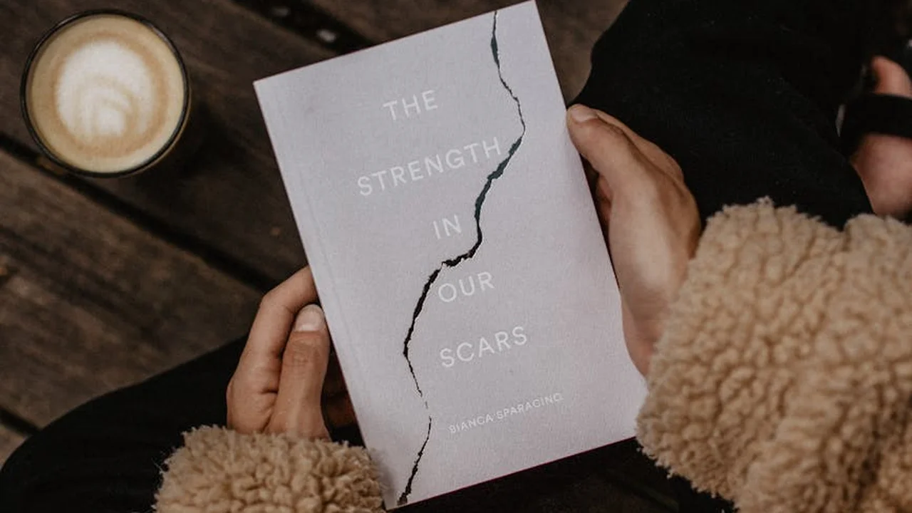 Close-up of a person holding a light-colored book titled 'The Strength in Our Scars' with a cracked cover, resting on a wooden surface beside a cup of coffee and a beige furry sleeve.