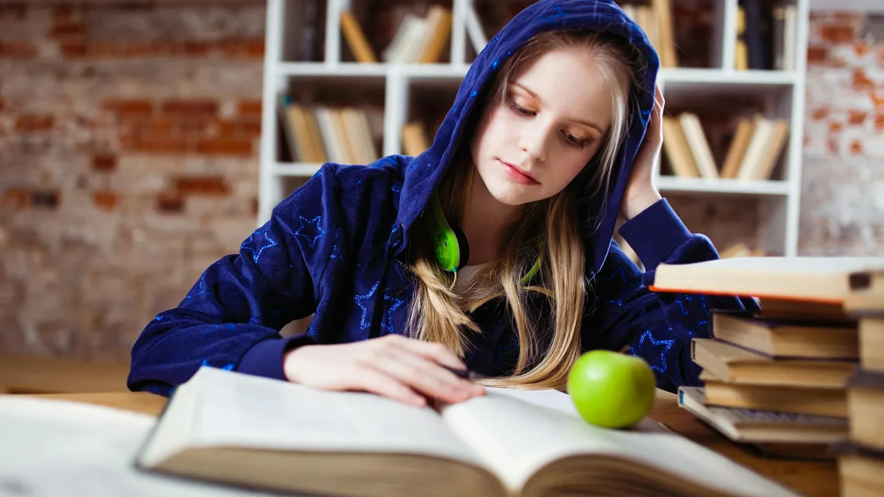 A young woman wearing a blue hoodie studies at a desk with an open book, a green apple, and a stack of books, in a library setting.