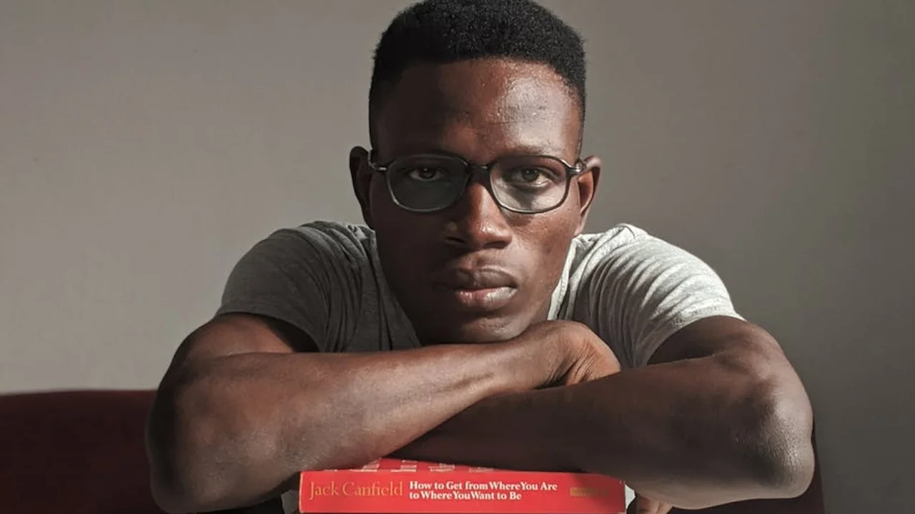 Portrait of a Black man wearing glasses, resting his arms on a table with a bold red book in the foreground.