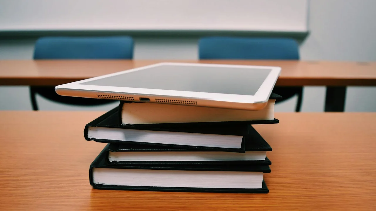 A tablet rests on a stack of books on a wooden desk, with blue chairs and a whiteboard in the classroom background.