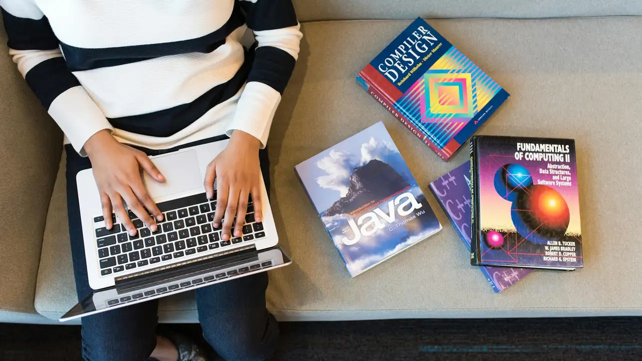 Person typing on a laptop with a stack of computer science books nearby, including Java and compiler design.