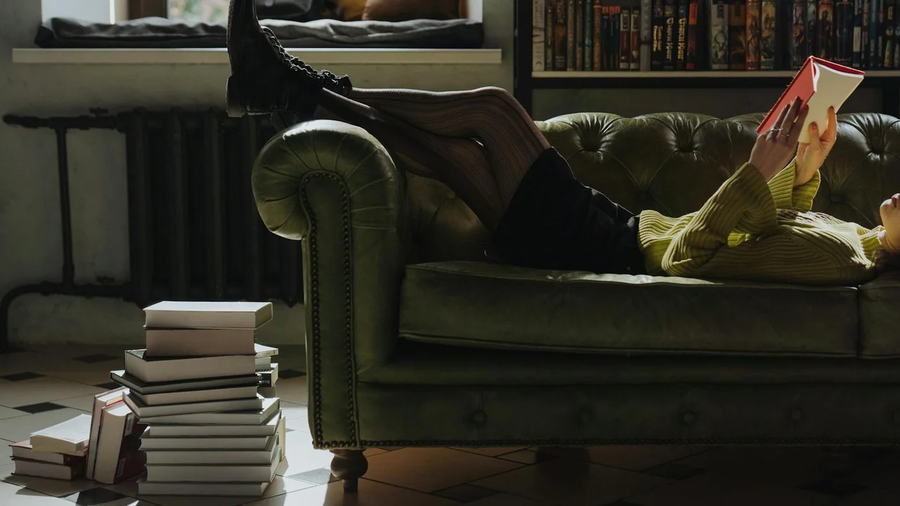 Person lounging on a green velvet sofa, wearing dark clothing and boots, reading a book with a stack of other books on the floor in a cozy living room
