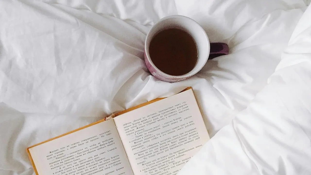 Open book on a white bed with a mug of tea nearby, evoking a calm, intimate reading moment for fans craving trauma, found-family, and deep emotional bonds.