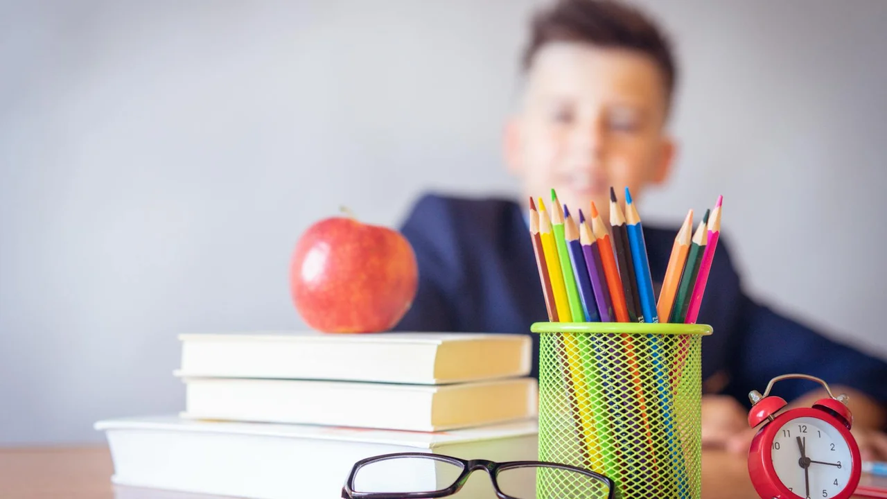 Blurred student in a classroom with an apple, a stack of books, and a cup of colored pencils on the desk.