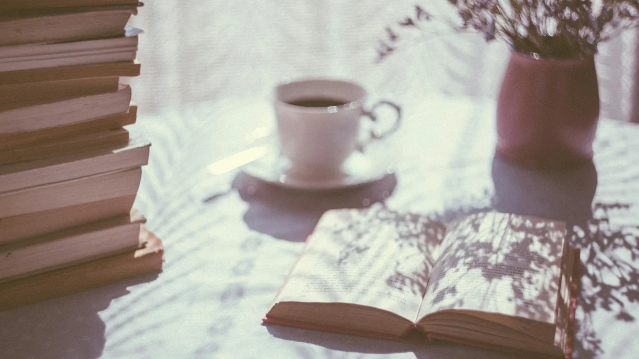 A desk setup featuring a stack of books on the left, an open book in the center, a small cup of coffee on a saucer behind it, and a vase of flowers on the right.