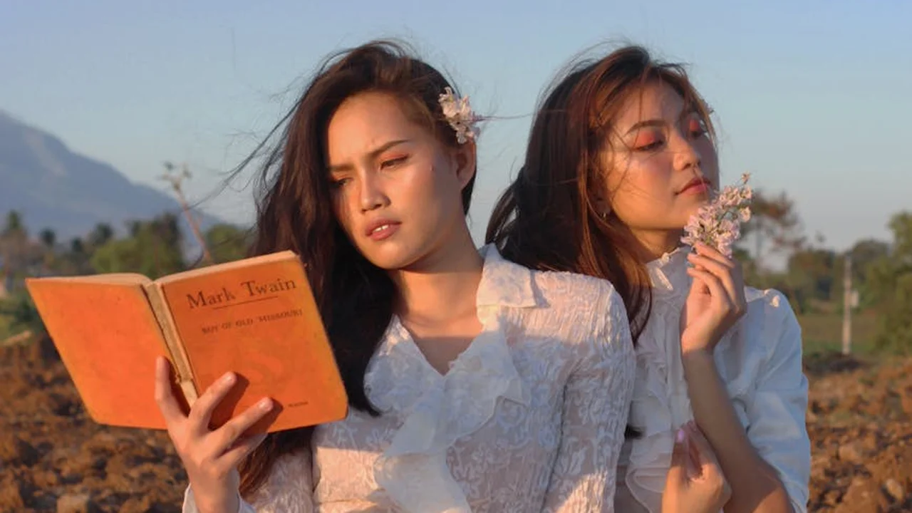 Two women in white dresses stand in a sunlit field. One reads an orange book titled Mark Twain, while the other holds a small flower to her face, with a rural landscape and distant mountains in the background.