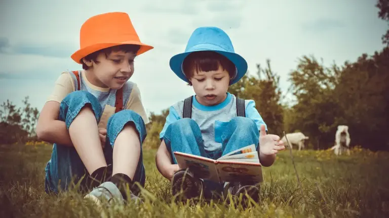 Two children sitting on grass outdoors, wearing bright orange and blue hats, reading a book together.