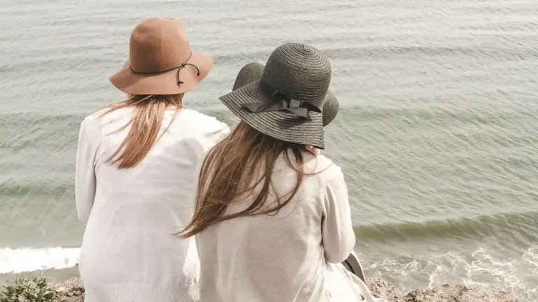 Two women wearing wide-brimmed hats sit at the water's edge, viewed from behind as waves roll onto the shore.