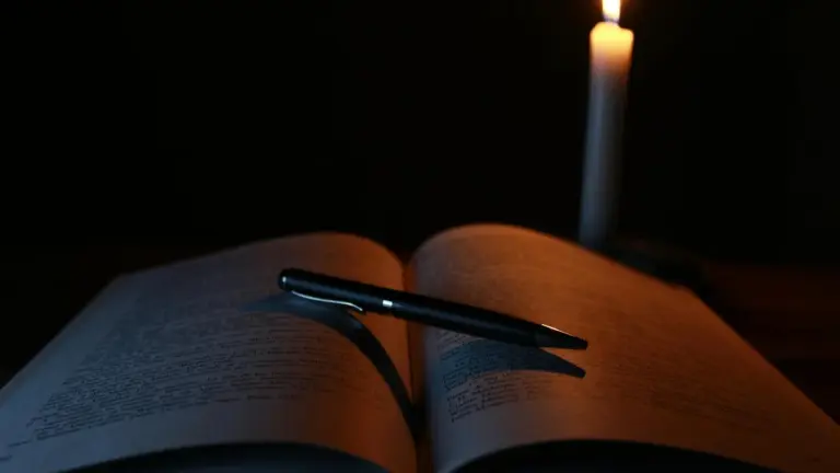 Open book with a pen resting on its pages, illuminated by a single candle in a dark room.